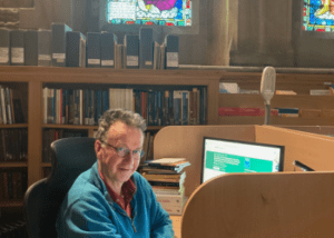 Angus sitting at his desk in OCMS surrounded by books and stained glass.